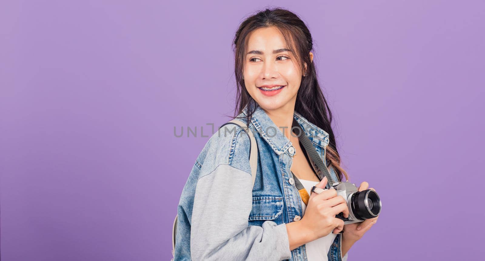 Attractive portrait happy Asian beautiful young woman smiling excited wear denims and bag holding vintage photo camera, female traveler female photographer, studio shot isolated on purple background