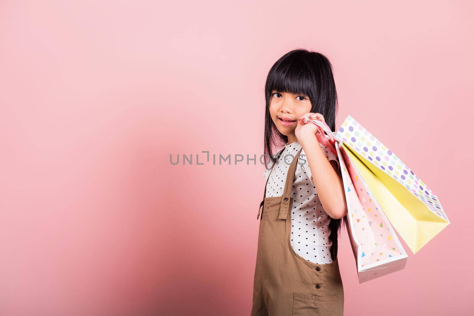 Asian little kid 10 years old smiling holding multicolor shopping bags in hands at studio shot isolated on pink background, Portrait of Happy child girl shopper lifestyle, Black friday concept