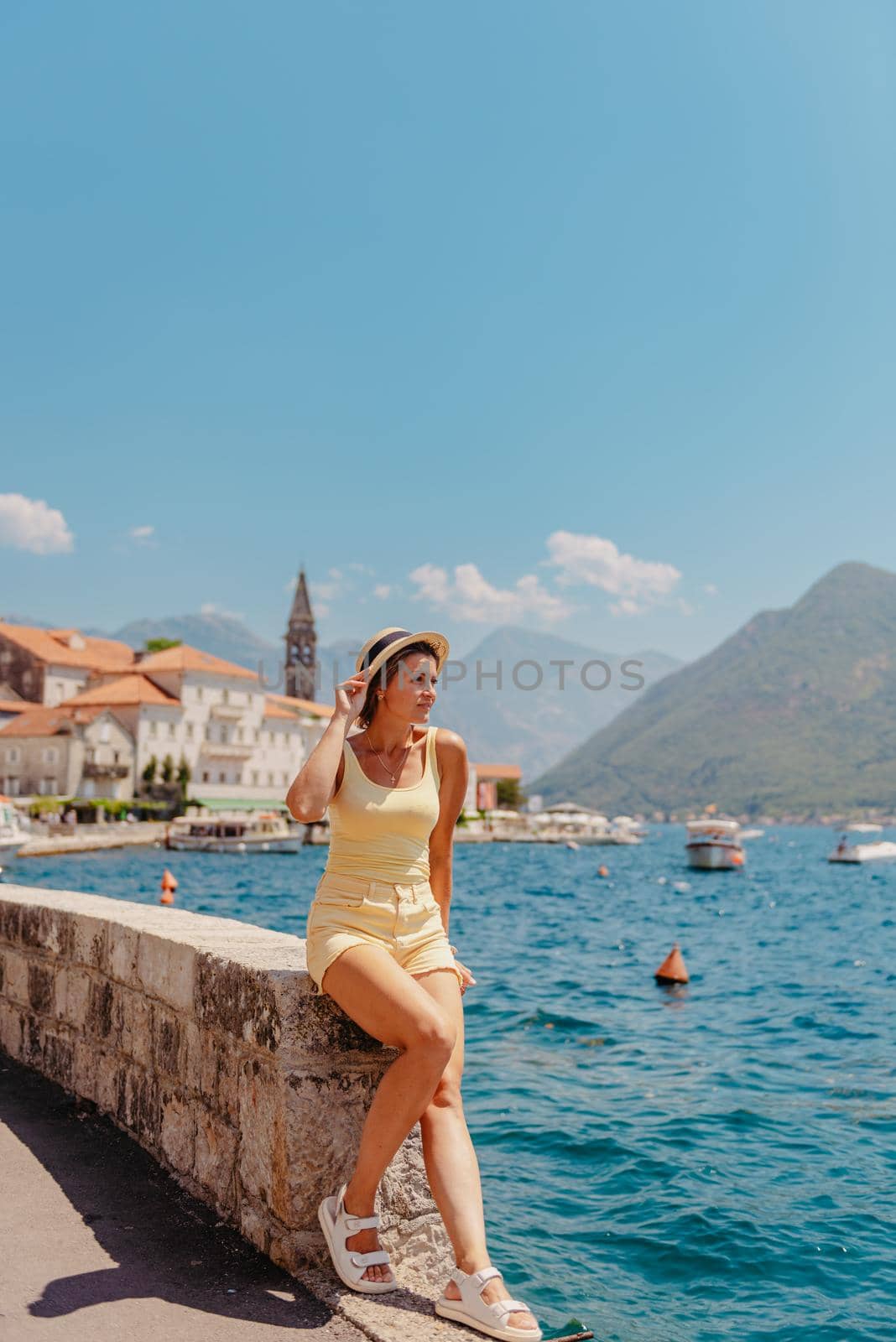 Summer photo shoot on the streets of Kotor, Montenegro. Beautiful girl in white dress and hat. smiling tourist girl with hat. Spectacular view of Montenegro with copy space. Ю fashion outdoor photo of beautiful sensual woman with blond hair in elegant dress and straw hat and bag, posing in Montenego's city Perast by Andrii_Ko