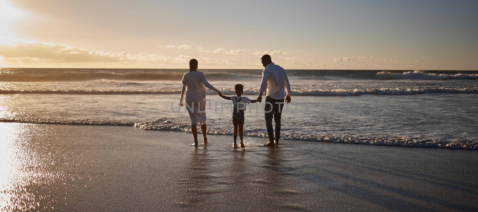 Rear view of a young african american family with one child getting their feet wet and having fun while spending time together by the beach at sunset.