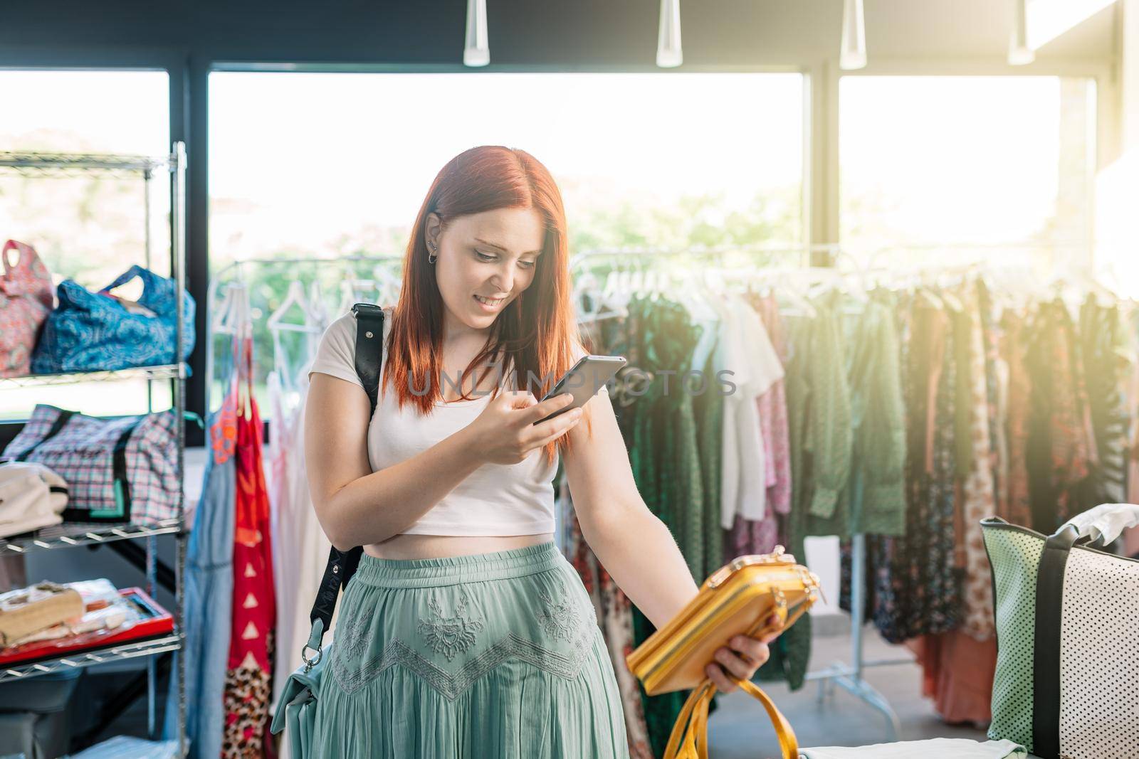 young red-haired woman happy shopping in a clothing shop, taking pictures with her smartphone of the clothes. girl spending time shopping sharing it on her social networks. concept of shopping. by CatPhotography