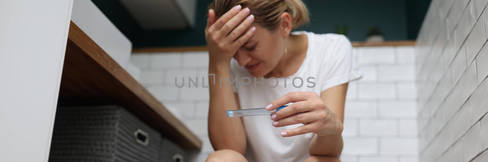 Low angle of young woman hold pregnancy test, got big news and feels upset about it. Blonde woman on toilet seat. Pregnancy, new period, changes concept