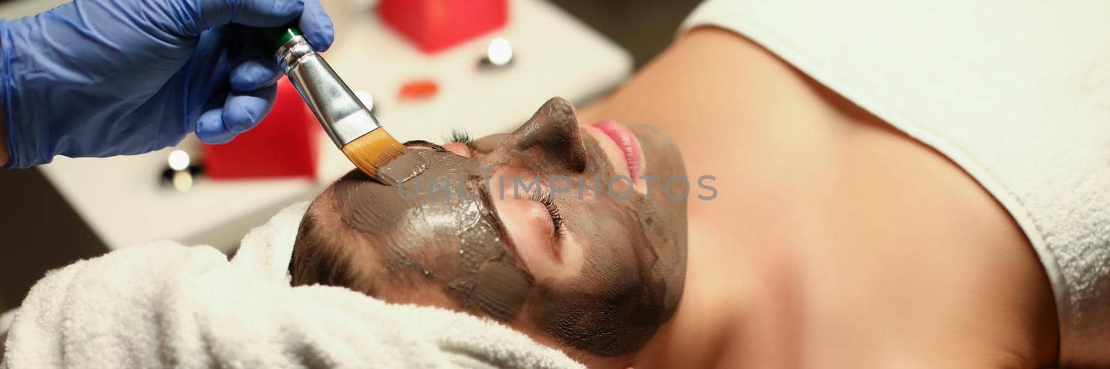 Top view of spa centre worker applying clay mask on clients face with special tool. Woman relaxing in cozy atmosphere. Wellness, skincare, beauty concept