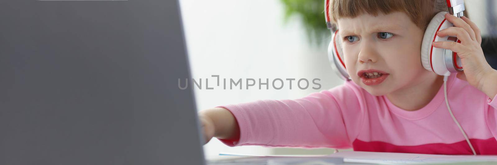 Portrait of little child not satisfied with something, point with finger on laptop screen. Upset kid in headset watch cartoon. Childhood, emotion concept