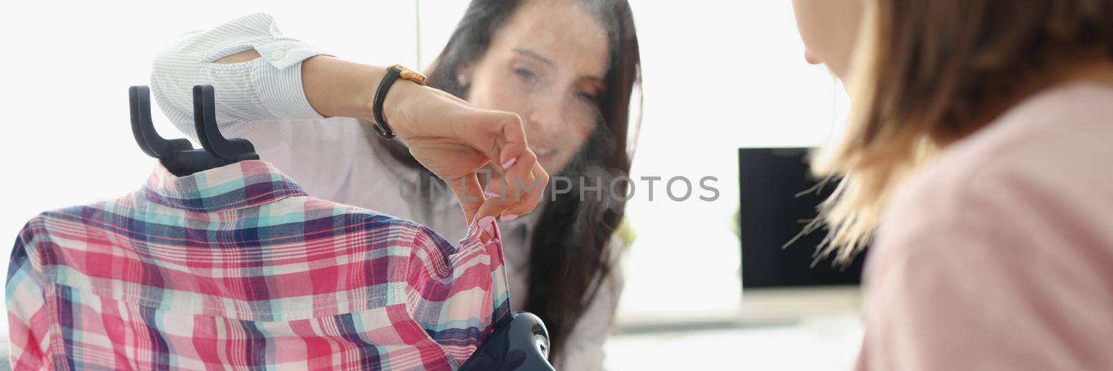 Portrait of girls from atelier use clothing steamer on plaid shirt, prepare product for selling. Cute women ironing glamour top. Couturier, sew concept