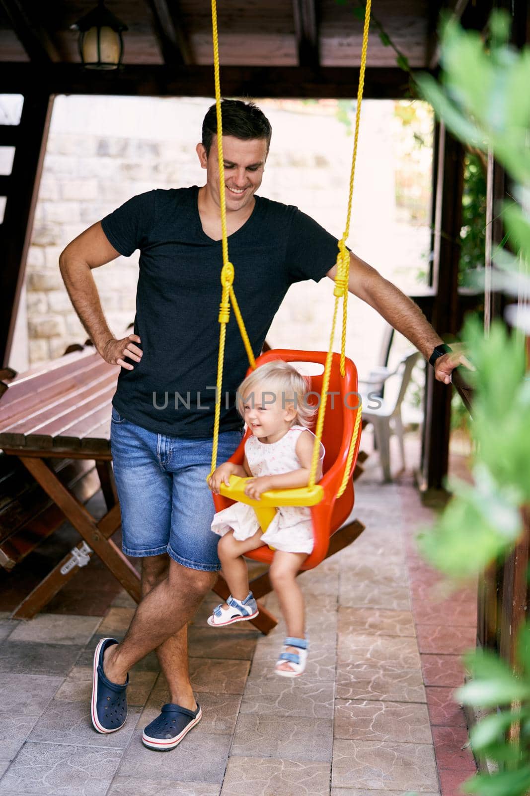 Smiling dad stands near a little girl on a swing in a wooden gazebo by Nadtochiy