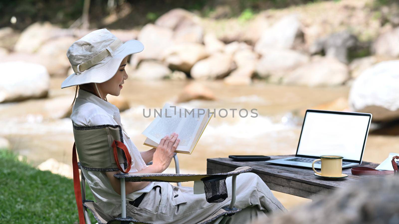 Calm female traveler reading book, enjoying camping alone near the river bank. Adventure, travel and camping concept.