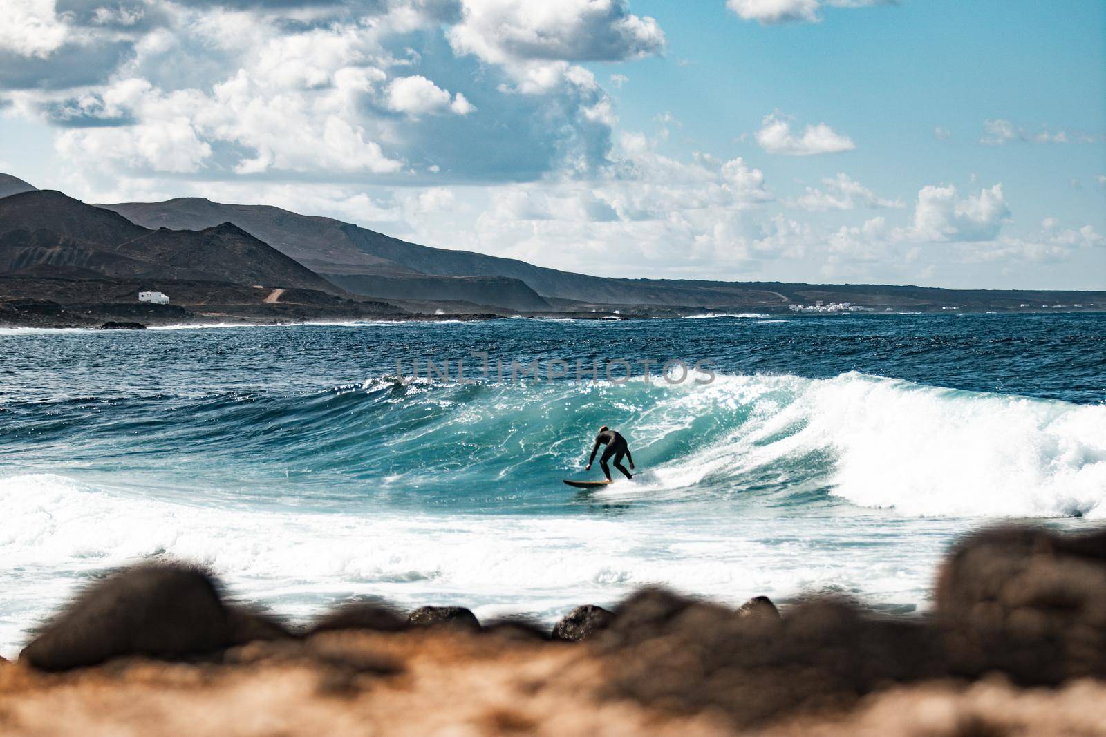 Wild rocky coastline of surf spot La Santa Lanzarote, Canary Islands, Spain. Surfer riding a big wave in rocky bay, volcano mountain in background