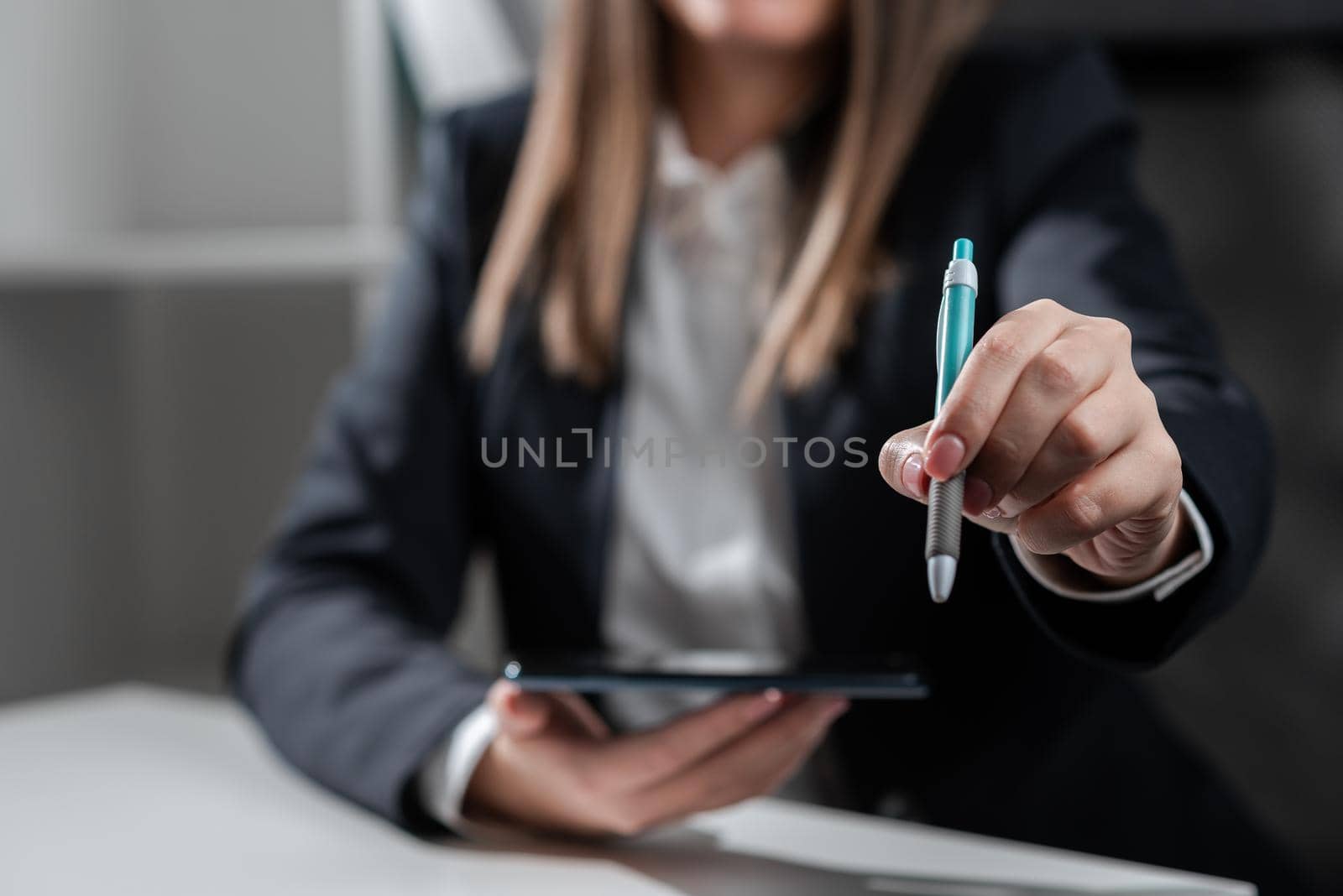 Businesswoman Holding Tablet In One Hand And Pointing With Pen On Important Message. Sitting Woman In Office With Cellphone In Hand Presenting Informations. by nialowwa