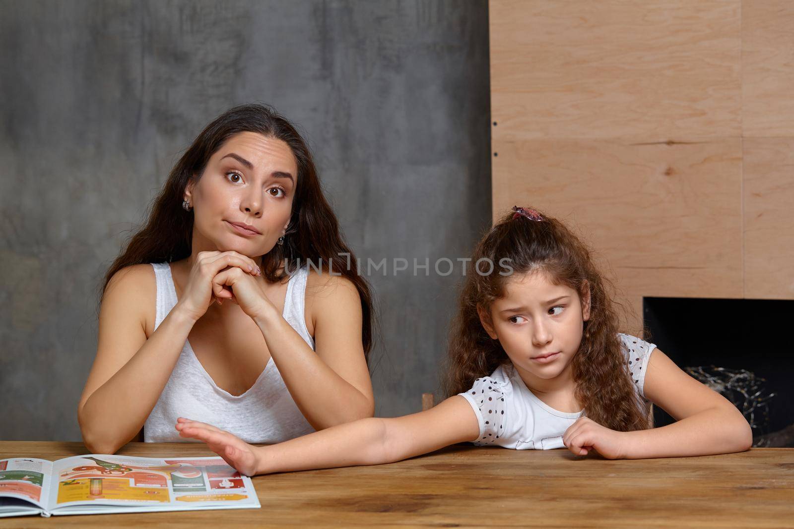 Portrait of a mother helping her small sweet and cute daughter to make her homework indoors. They both are disappointed. Mom is looking at the camera and her daughter is looking away.