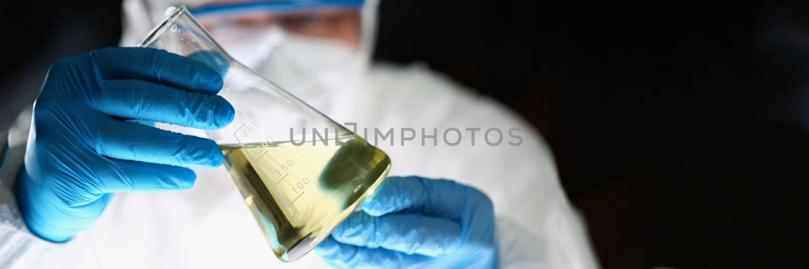Close-up of laboratory pharmacist in respirator and uniform show flask with yellow liquid. Chemist work on test in blue gloves. Chemistry, science concept