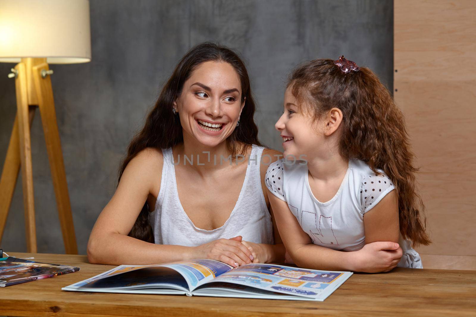 Portrait of a mother helping her small and cute daughter to make her homework indoors. They are looking at each other and smiling.