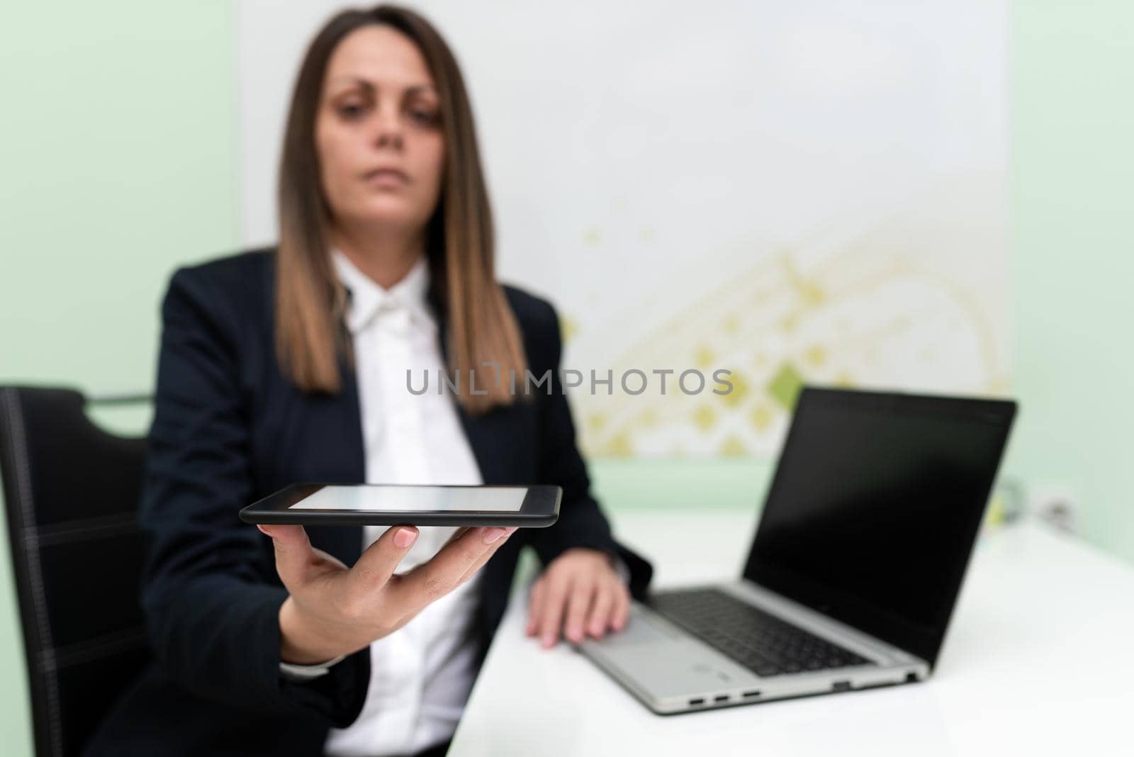 Businesswoman Holding Tablet With One Hand And Having Lap Top On Desk. Woman Presenting Important Data On Phone Screen. Sitting Executive On Desk Showing New Ideas. by nialowwa