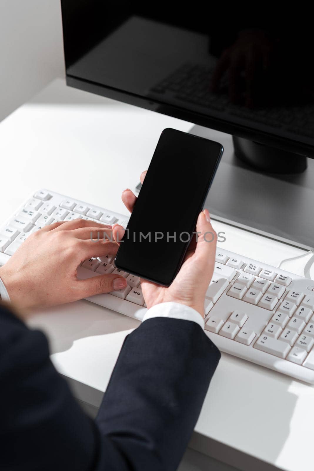 Businesswoman Holding Mobile Phone With Important Announcements Sitting On Desk With Computer. Woman In Suit Having Cellphone With Crutial Informations. by nialowwa