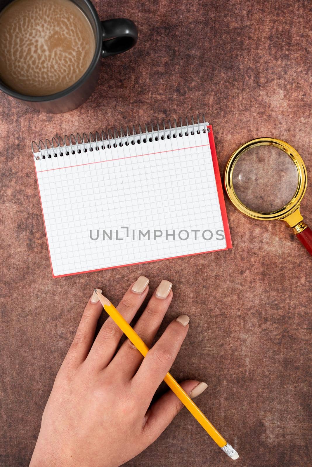 Hand Of Woman Having Spiral Notebook With Copy Space, Pencil, Magnifying Glass And Coffee Cup Over Table. Businesswoman Writing Important Message On Book With Drink. by nialowwa