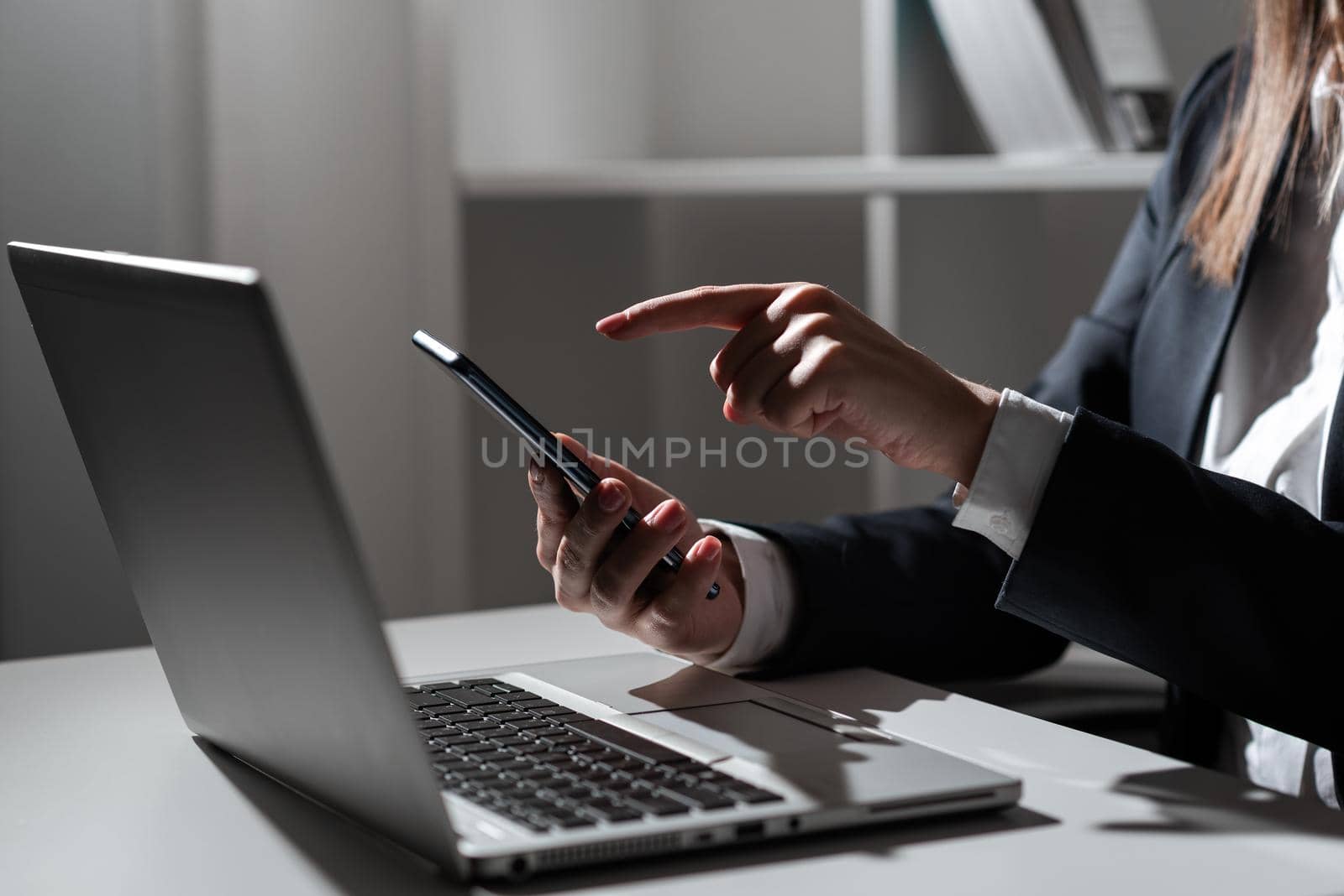 Businesswoman Holding Tablet In One Hand And Pointing On Important News With One Finger. Sitting Woman In Office With Cellphone In Hand Presenting Informations. by nialowwa