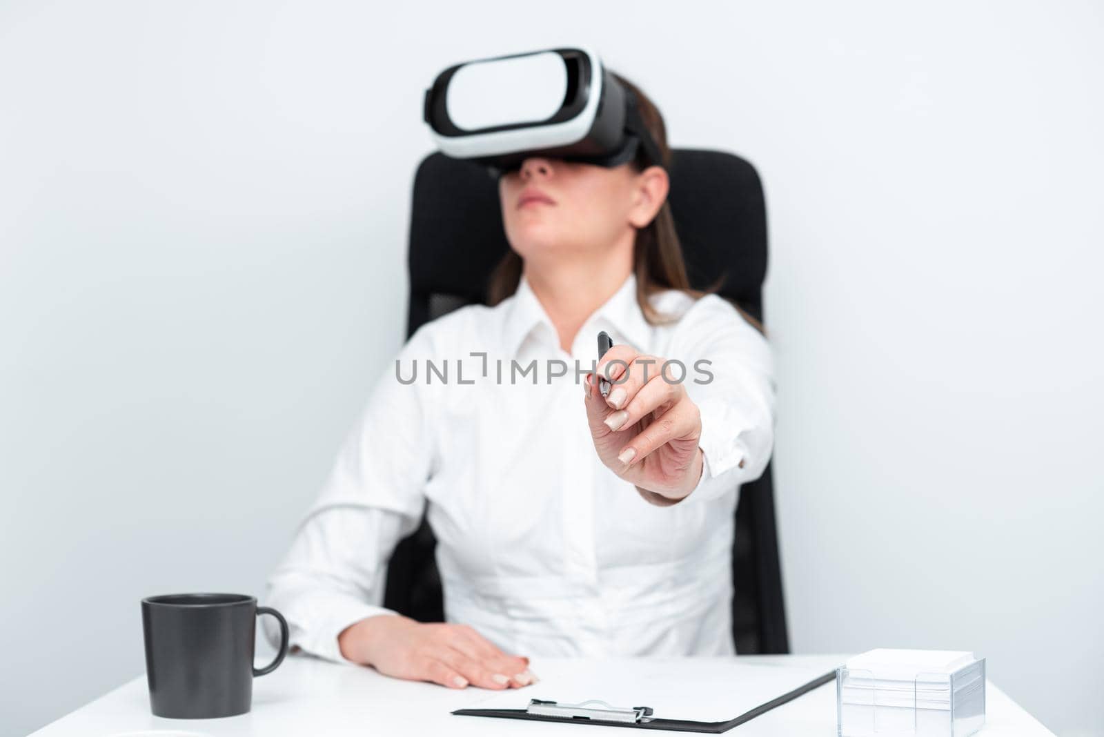 Businesswoman Wearing Virtual Reality Goggles And Learning Professional Skill Through Futuristic Simulation. Woman Holding Pen And Sitting At Desk During Modern Training. by nialowwa