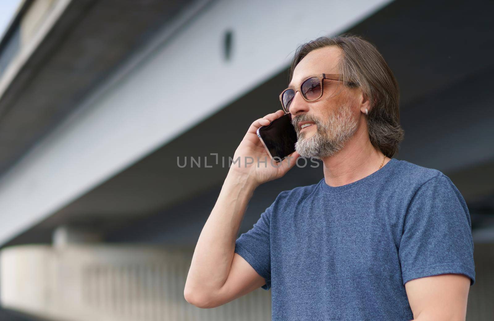 Middle aged using phone in city. Man on urban background talking on the phone. Travel concept. Handsome stylish sexy middle aged grey bearded man in sunglasses under the bridge wearing casual.