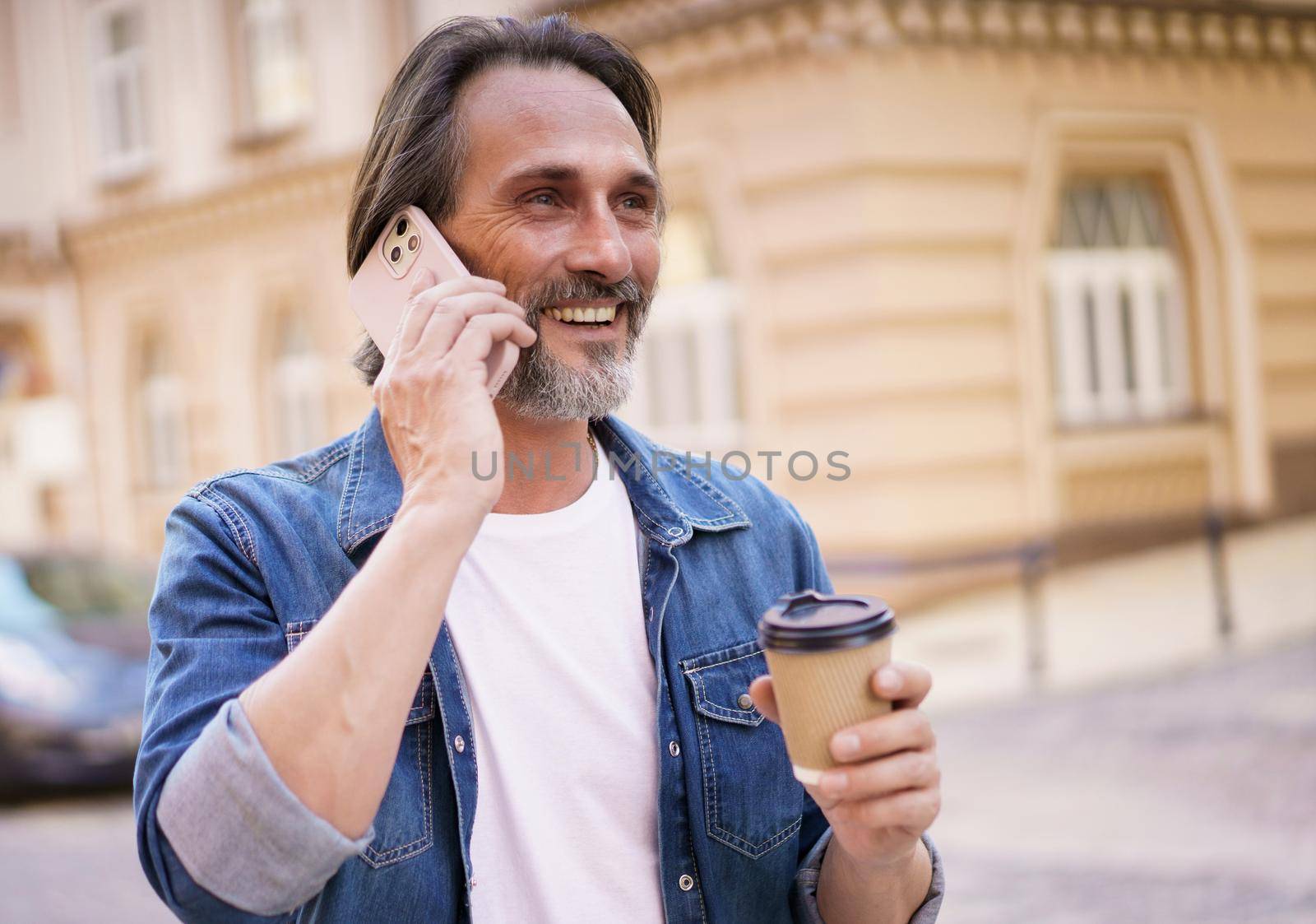Happy middle aged man with grey bearded talking on the phone holding coffee in disposable paper cup standing outdoors in old city background wearing jeans shirt. Freelancer traveling man.