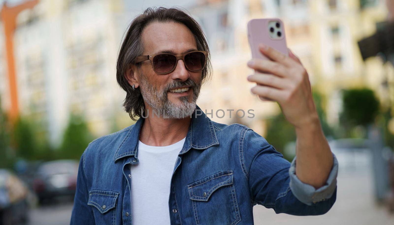 Handsome stylish sexy middle aged grey bearded man smiling on camera old town wearing casual. Happy senior man on street of european city making selfie using mobile phone. Travel concept by LipikStockMedia