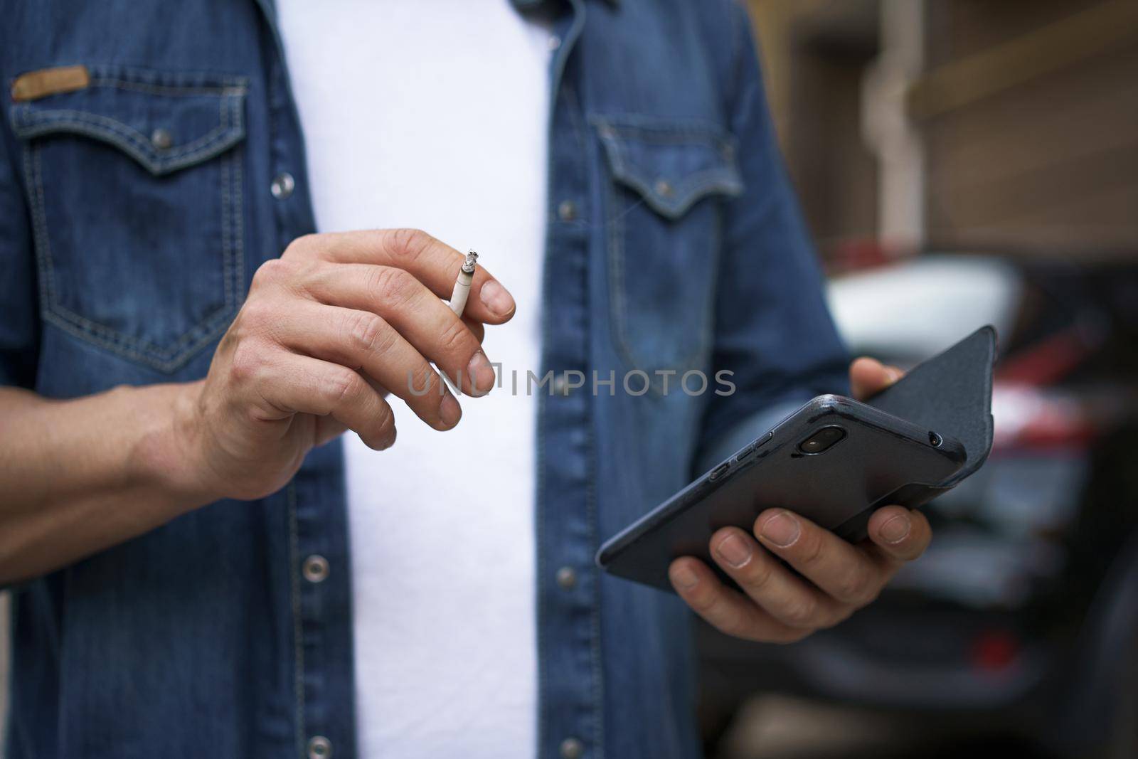 Close up. Unhealthy addiction smoking cigaret man read social media using his smartphone wearing jeans shirt and white t-shirt standing outdoors on urban city background. No face visible.