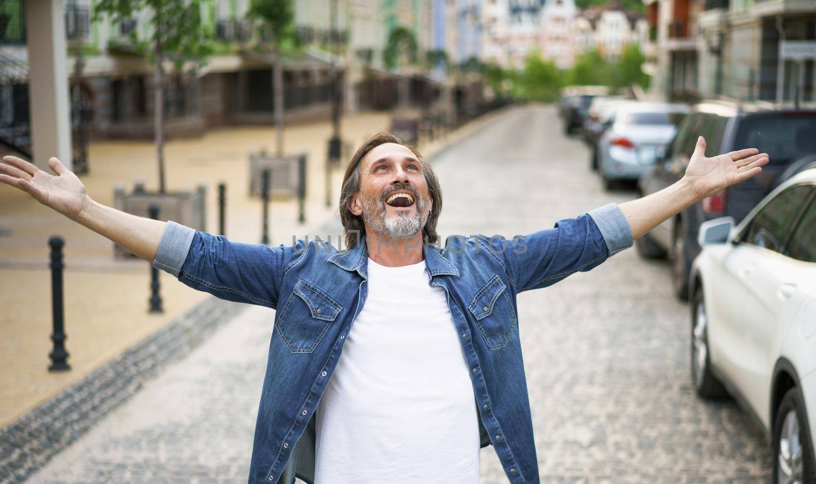 Happy senior man thanks God standing outdoors with hands spread wide open wearing jeans shirt with white t-shirt under. Mid Aged man glad that life is beautiful standing in old town street.