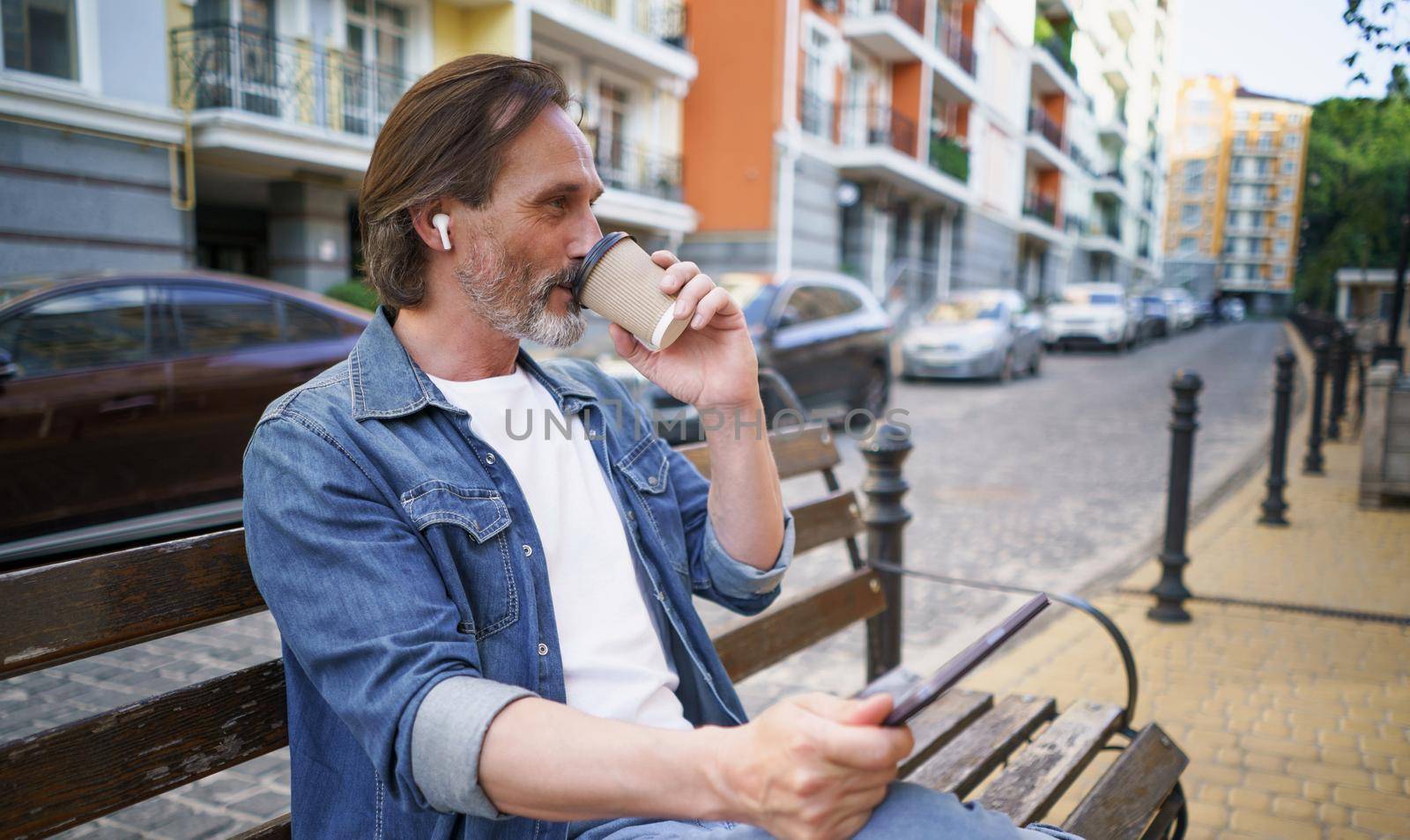 Sitting alone on the bench middle aged man away of office holding digital tablet, wireless earphones having a call drinking take away coffee at old town streets. Business on the go concept by LipikStockMedia