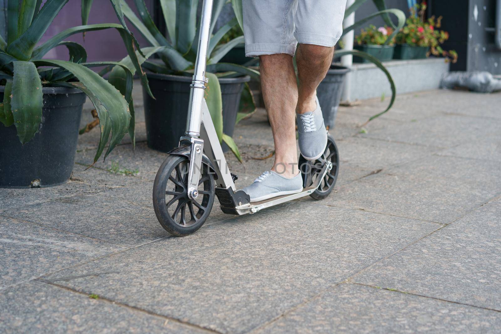 Close up male feet in sneakers shoes stand on scooter with big wheels having a ride on the streets or park after work outdoors with agave plants on background. No face visible. Focus on front wheel.