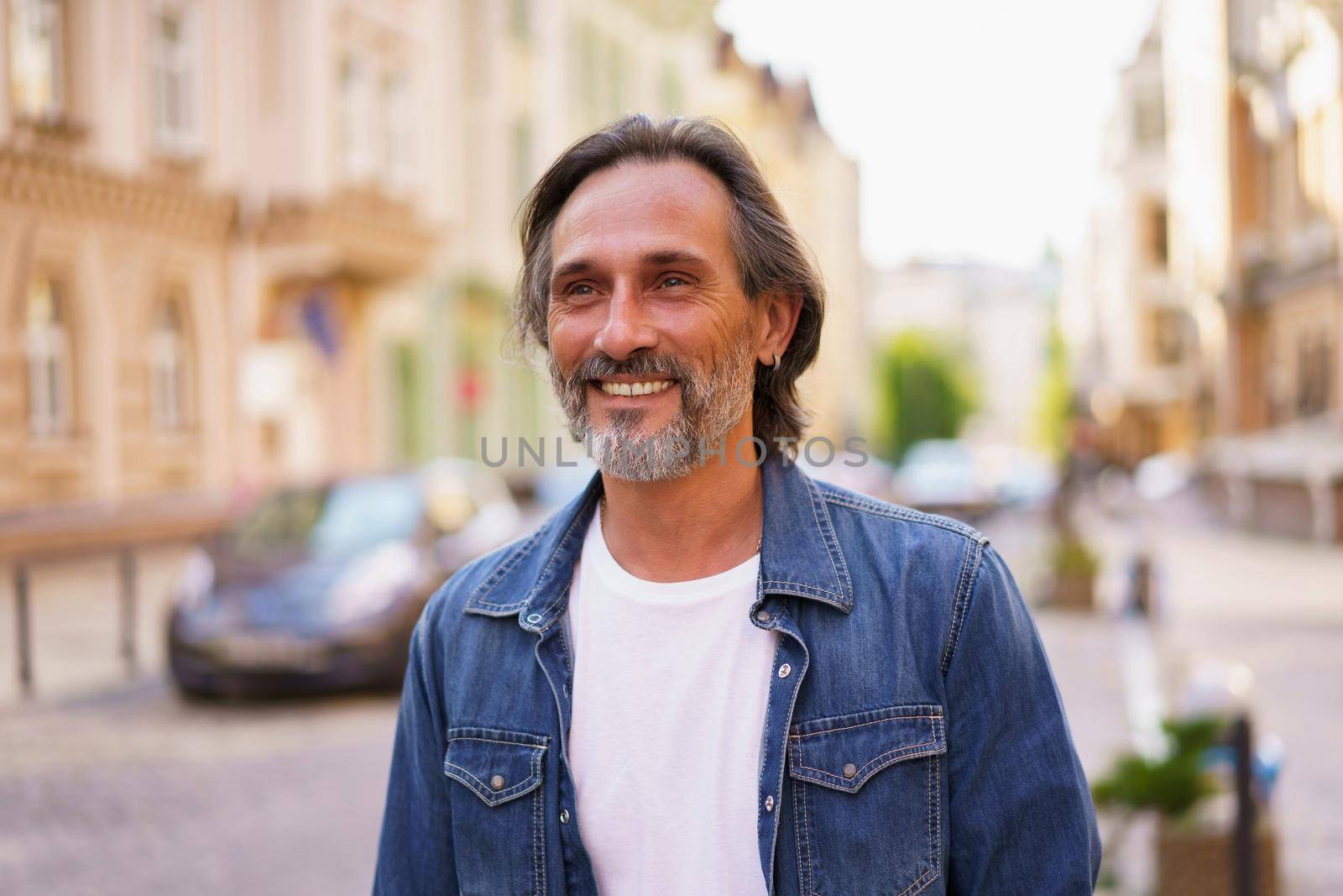 Well dressed happy mature man outdoors in old town city smiling wearing jeans shirt and white t-shirt. Happy mature man wearing sunglasses and looking sideways outdoor on a summer day.