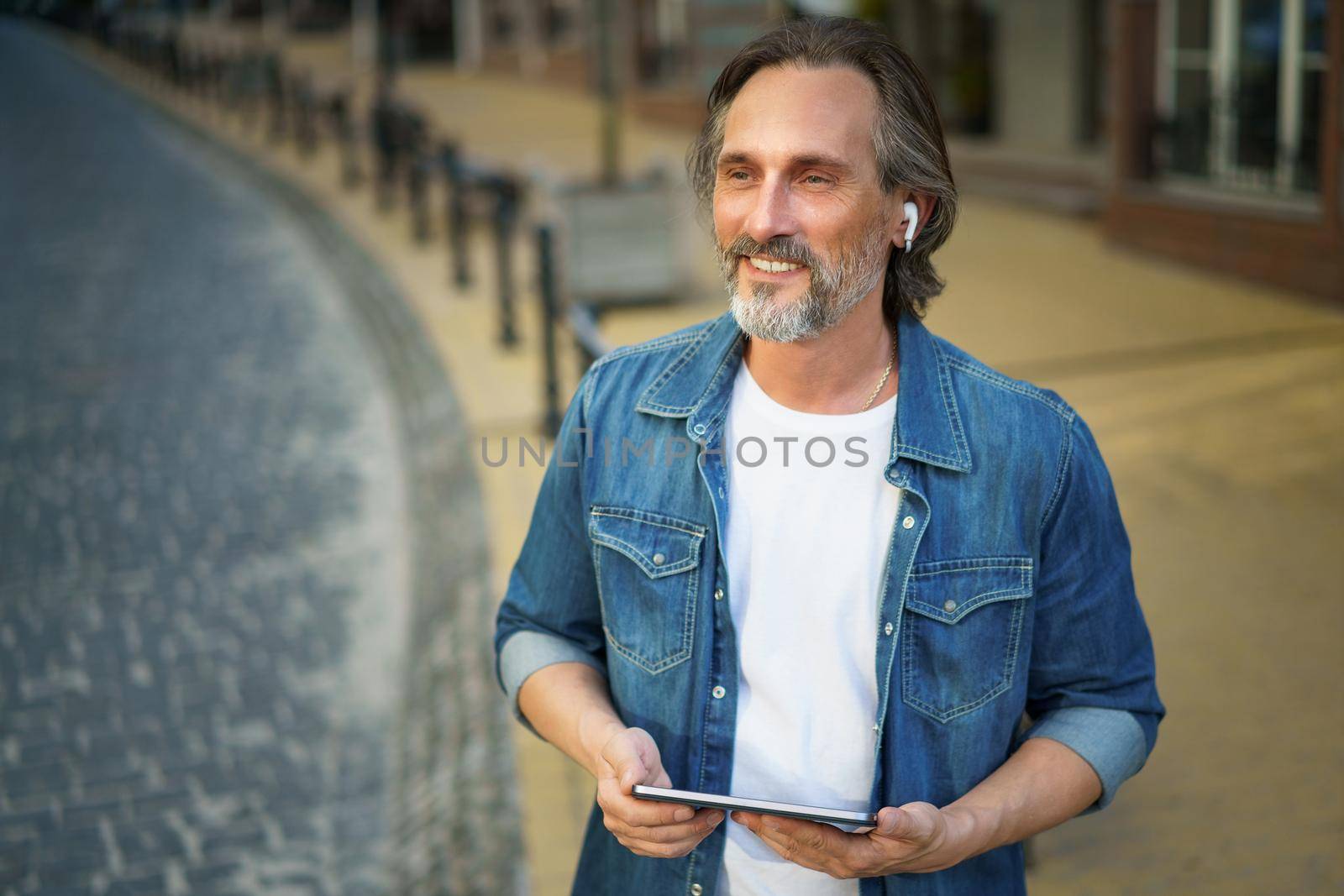 Traveling handsome middle aged man using digital tablet having a call while standing on urban city streets. Mature man talking or listening music use wireless earphones while travel old town streets.