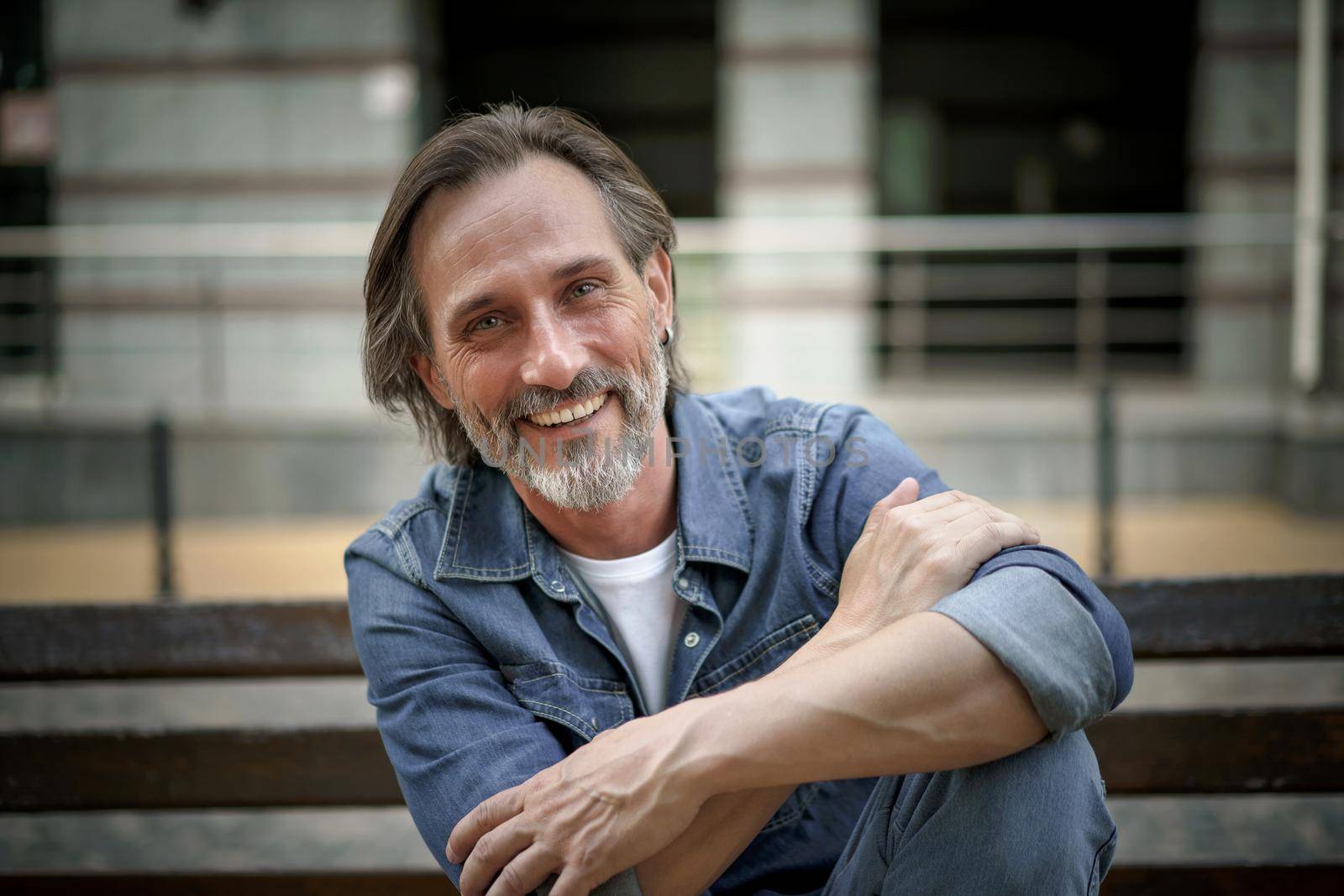 Smiling handsome middle aged man with grey beard sits on the bench with hands folded leaned on one lifted foot outdoor at urban city background enjoying free time. Travel and people concept.