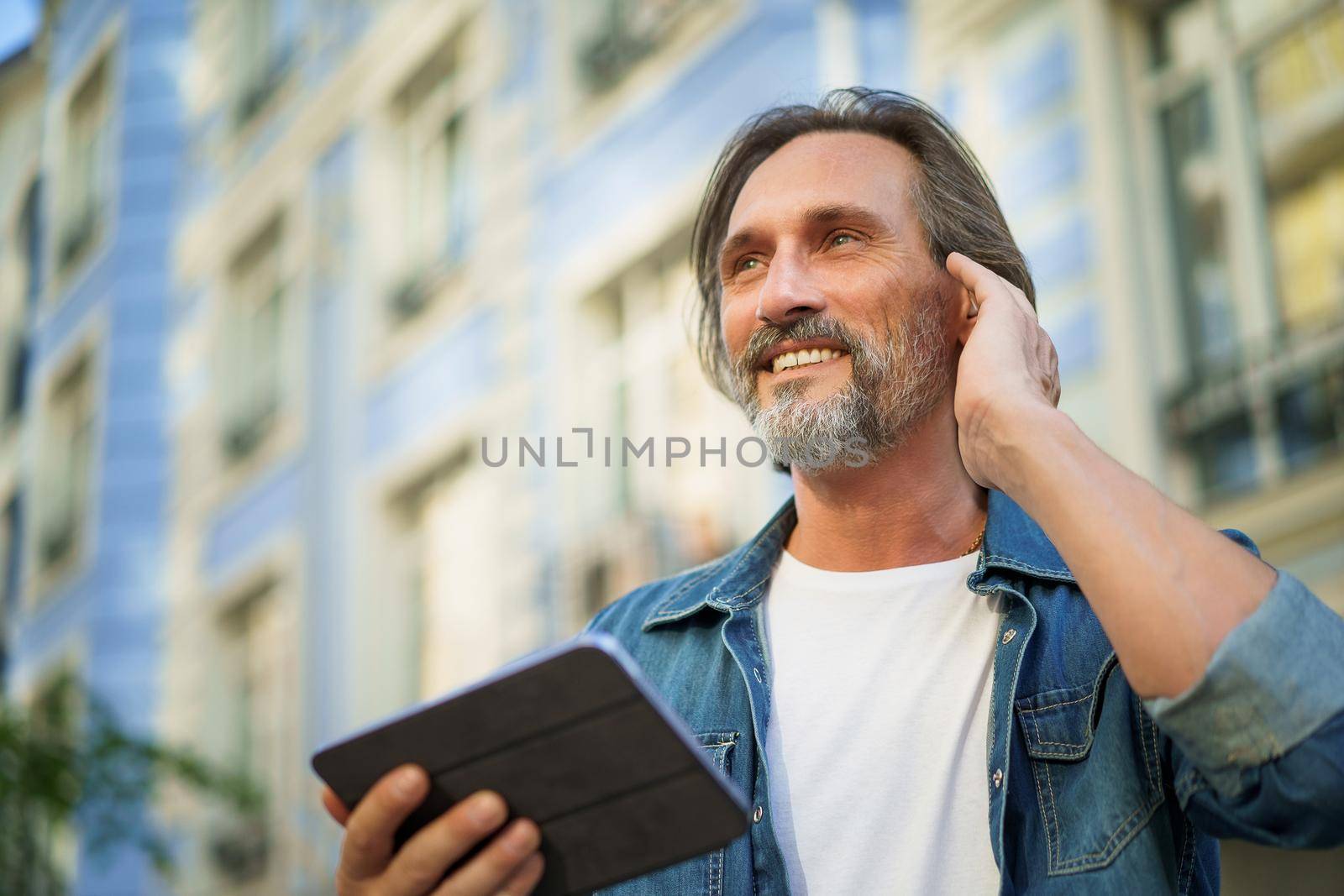 Putting in wireless earphone handsome traveling mature man use digital tablet talking outdoors having a call on urban city streets. Mature man listening music travel old town streets.