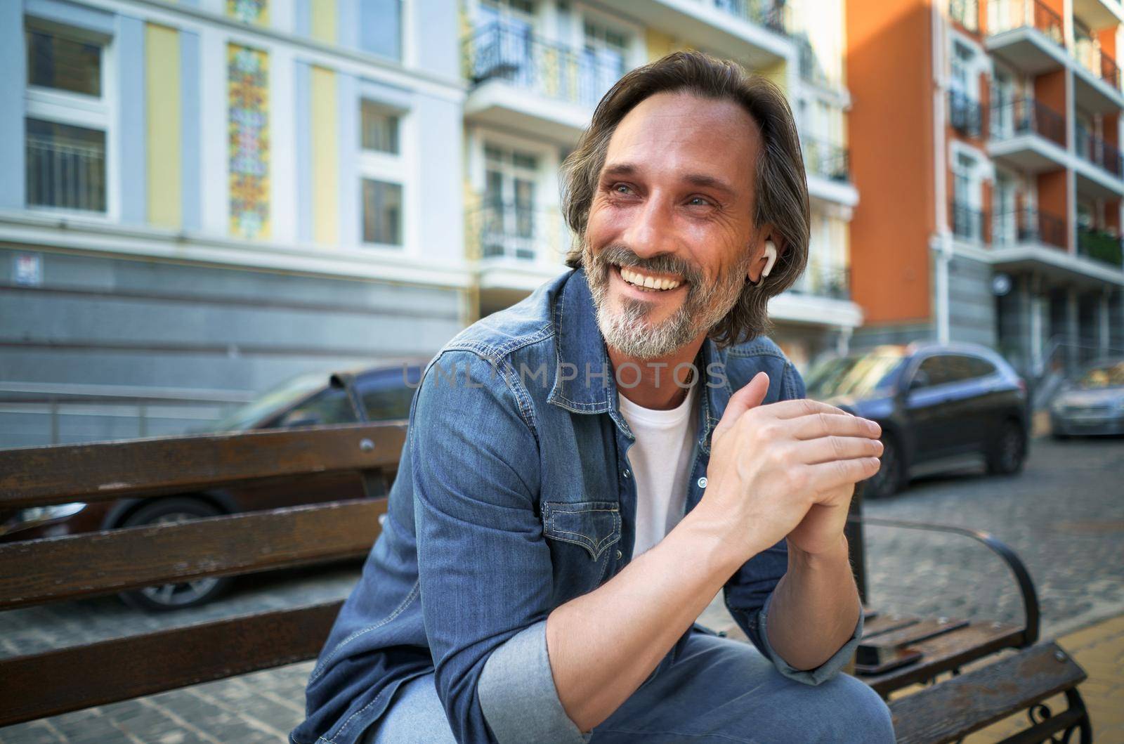 Smiling positive emotion middle aged man leaned his hands on knee looking sideways sitting at the bench in old town. Happy mature grey bearded man with positive emotions on his face.