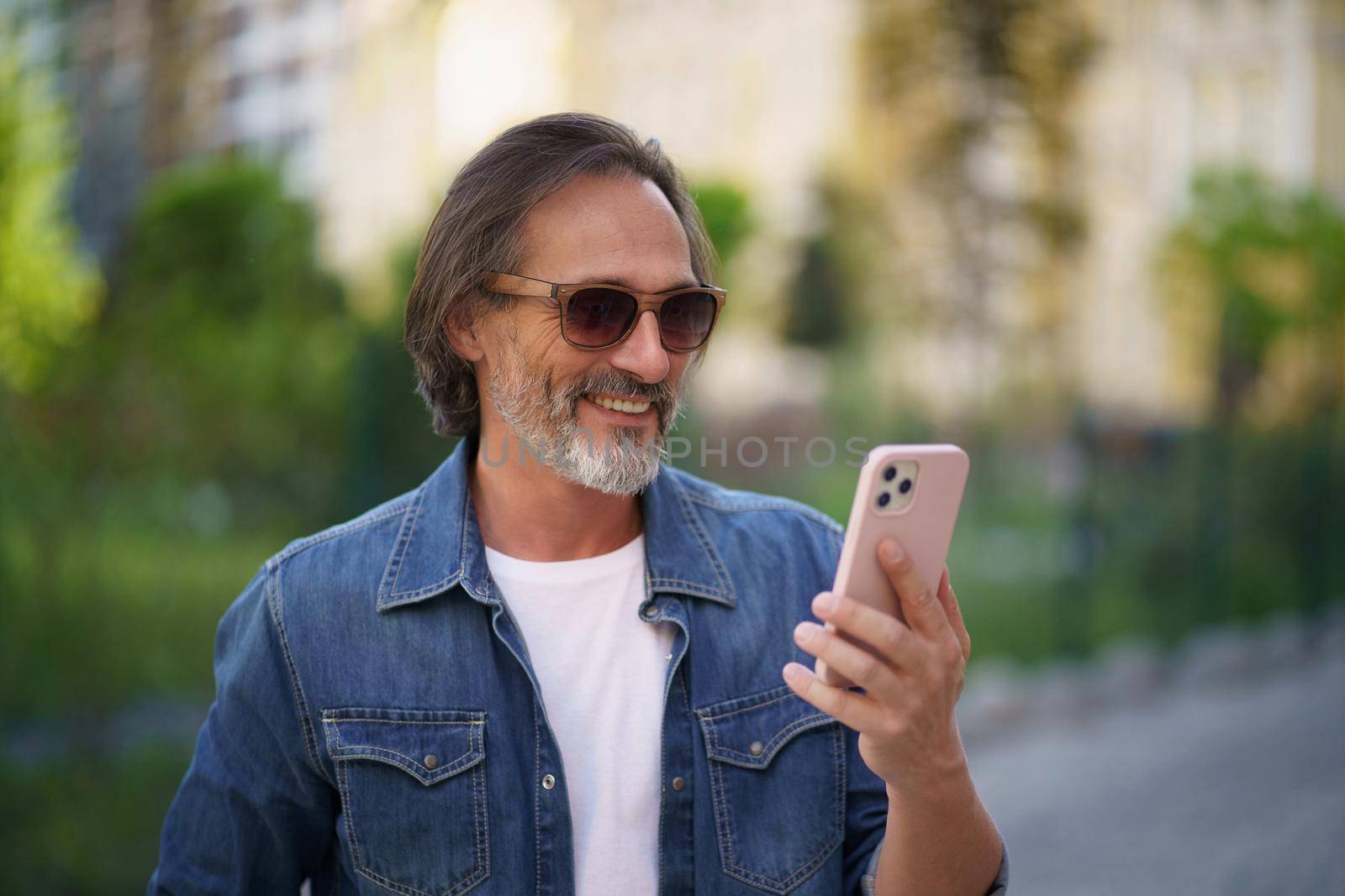 Happy adult man on street of european city making selfie using mobile phone. Travel concept. Handsome stylish sexy middle aged grey bearded man smiling on camera old town wearing casual by LipikStockMedia