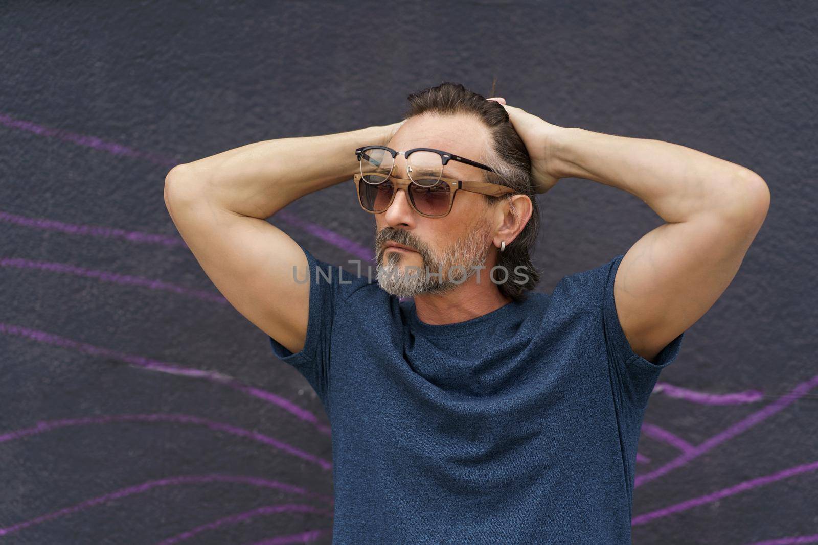 Middle aged man put hands behind head with glasses and sunglasses on his face standing next to dark urban wall wearing dark blue t-shirt. Mature business man or freelancer outdoor.