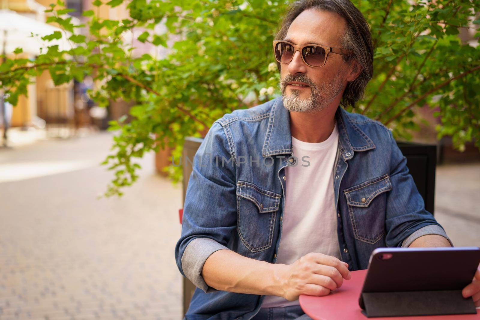 Handsome mature freelancer man enjoying coffee outdoors while having group chat. Middle aged man looking away sitting at cafe or street restaurant while working outdoors using digital tablet.