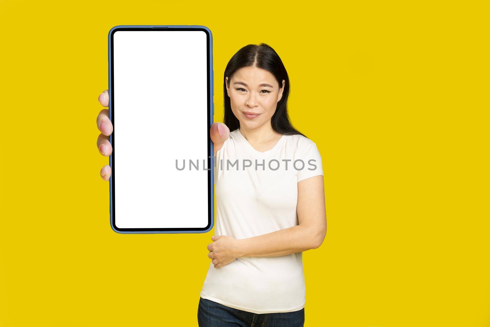 Showing phone white screen middle aged asian woman 40s holding smartphone mobile app advertisement and excited smile on camera isolated on yellow background. Product placement by LipikStockMedia