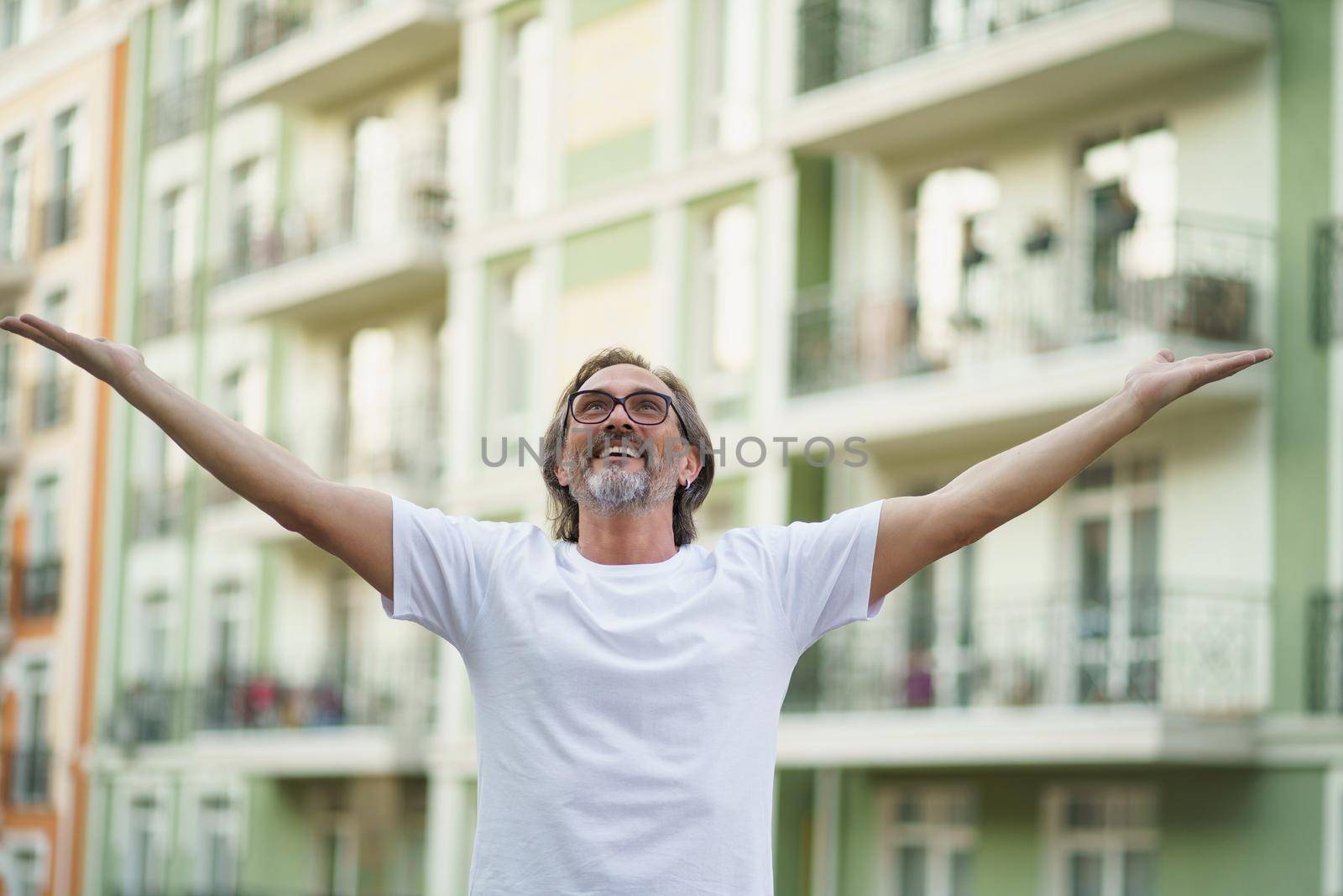 Middle aged man spread his hands in the air praising God standing outdoors wearing white t-shirt and eye glasses. Middle aged man glad that life is beautiful standing in old town street.