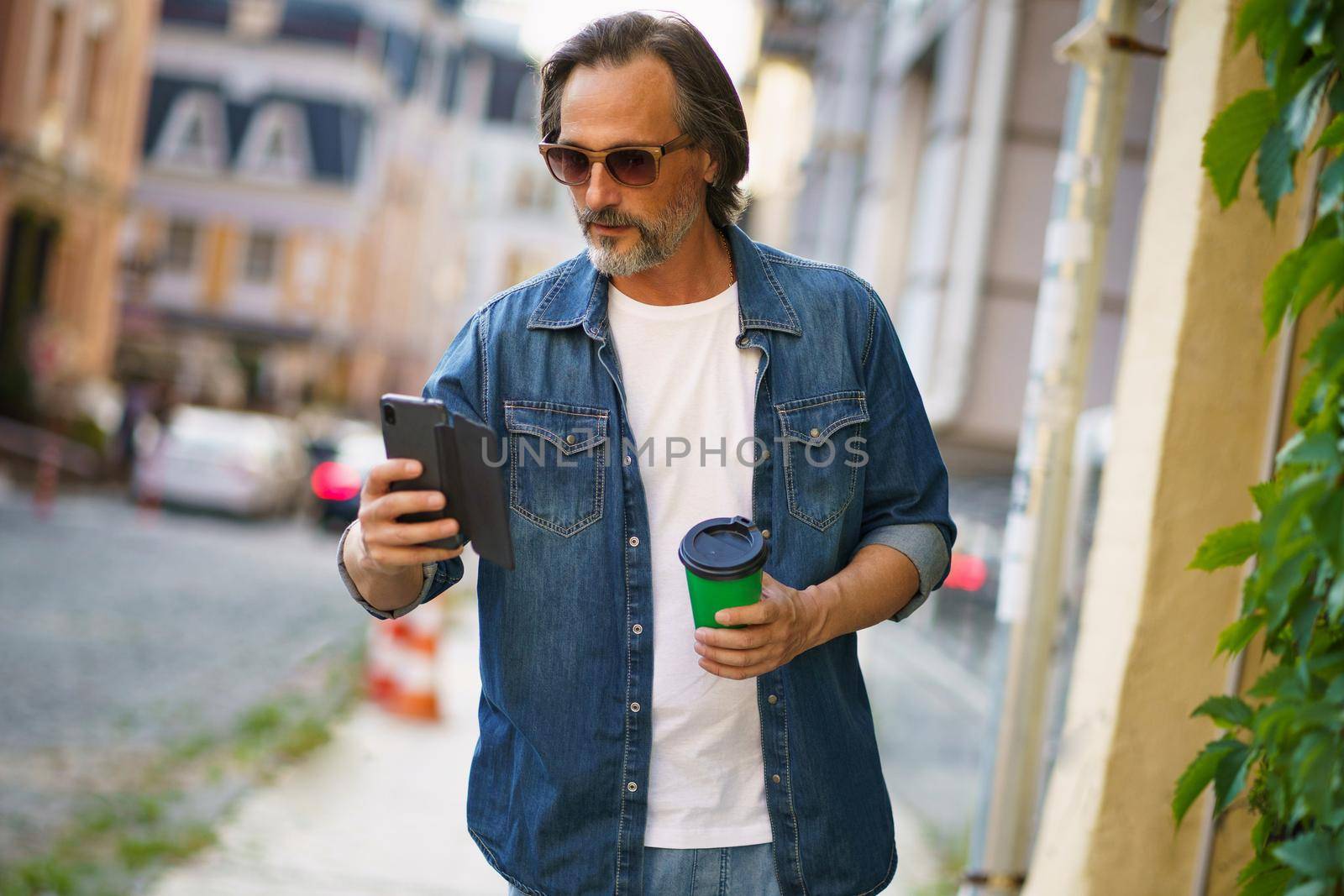 Mature man read text or email walking off work or back to office holding phone and take away paper cup with hot coffee wearing sunglasses with at old town streets. Business on the go concept by LipikStockMedia