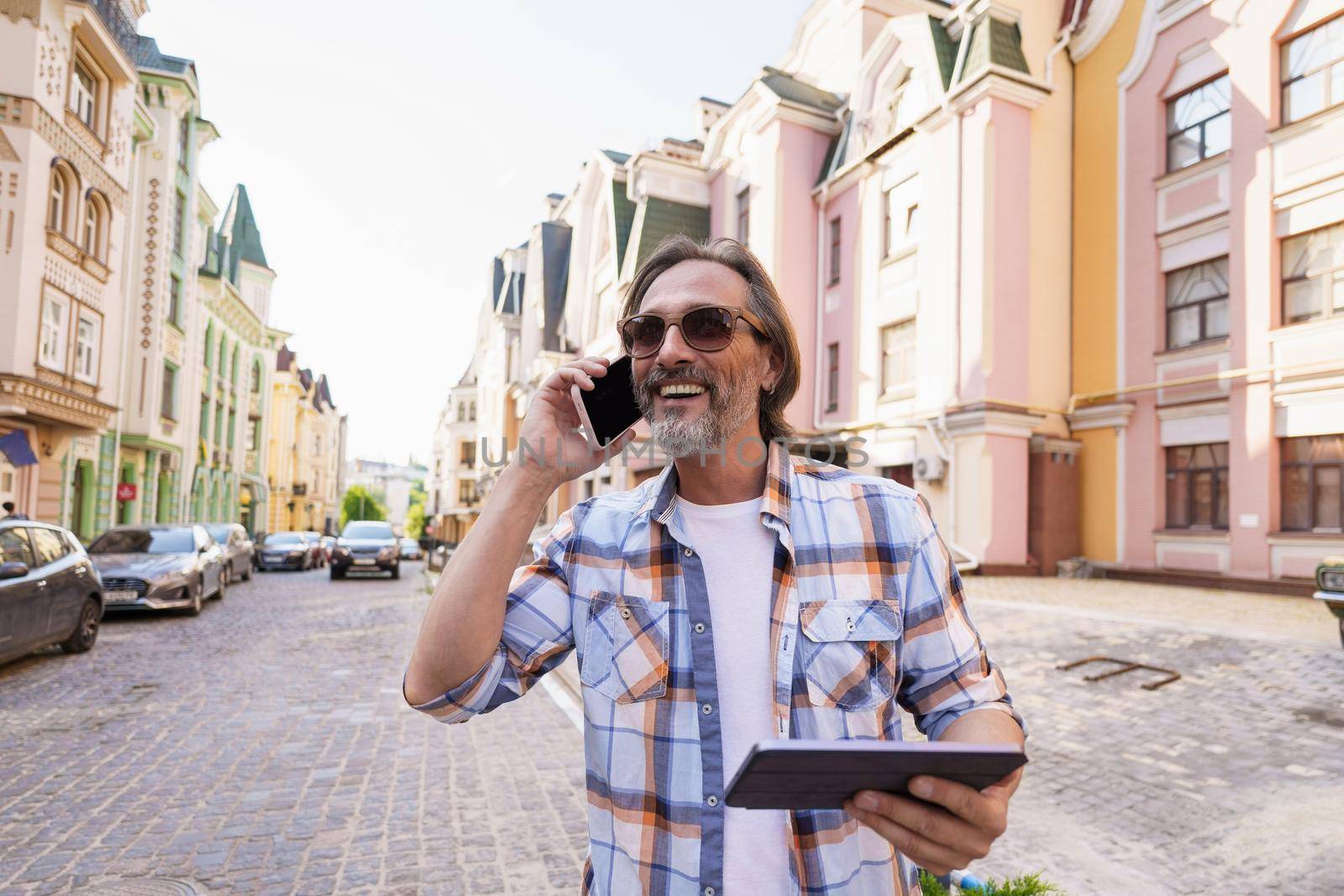 Happy smiling middle aged man with grey beard talking on the phone holding digital tablet in hand standing outdoors in old city background wearing plaid shirt and sunglasses. Freelancer traveling man.