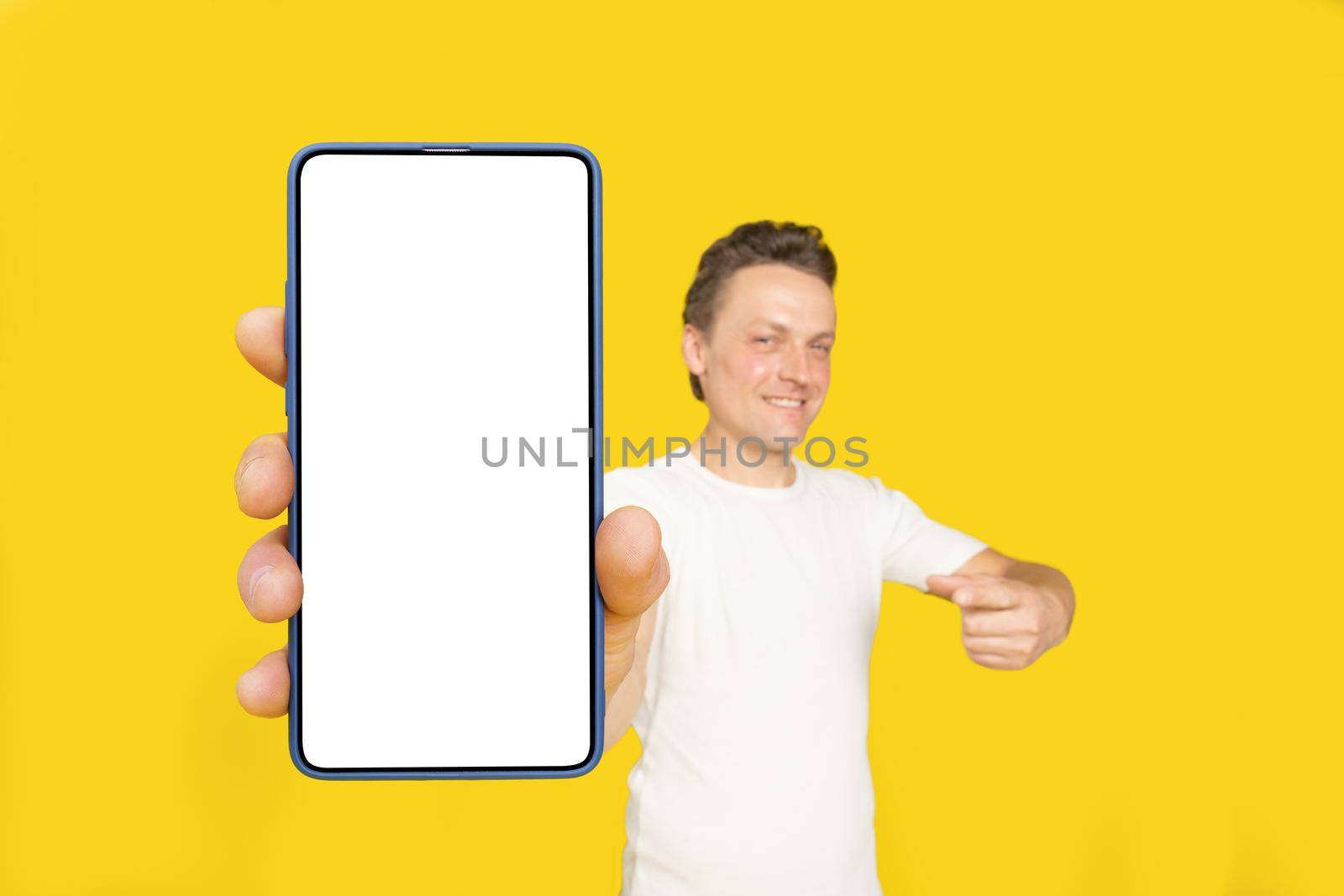 Huge smartphone in hand of handsome blond man pointing at white empty screen, wearing white t-shirt and jeans isolated on yellow background. Mobile app advertisement, great offer.