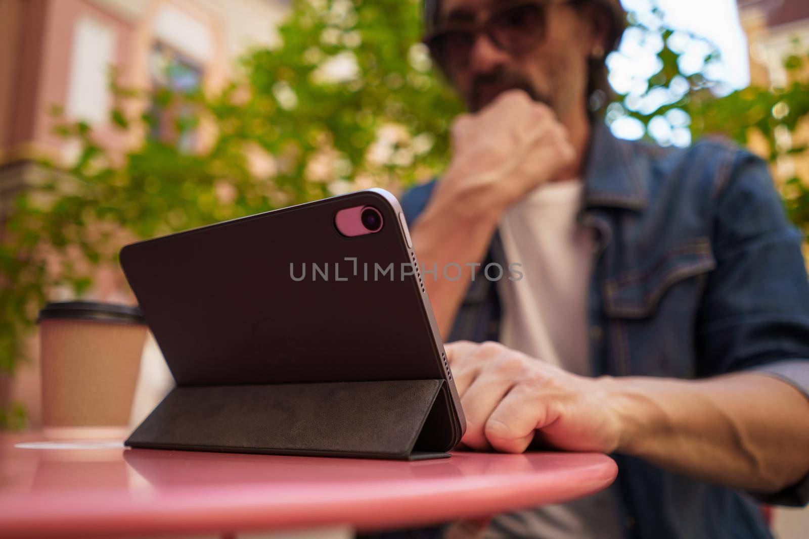 Low view middle aged man working outdoors using digital tablet drinking coffee. Handsome freelancer traveling man checking work or social media sitting at city street cafe. Selective focus on tablet.