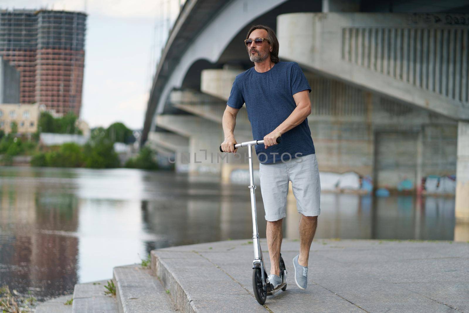Riding scooter handsome stylish middle aged man with grey beard stand under town bridge over river with urban city on background after work outdoors. Travel, lifestyle concept.
