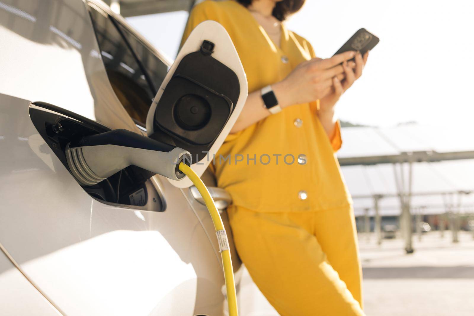 Woman stands near recharging electric car with mobile phone. Vehicle charging at public charging station. Electric car charging near an solar power plant.