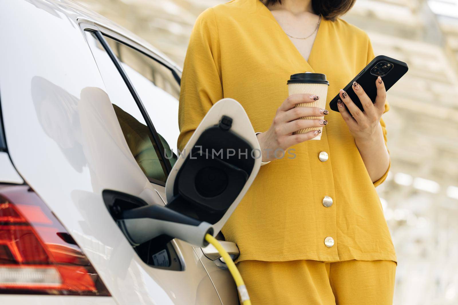 Unrecognizable caucasian woman using smart phone and waiting power supply connect to electric vehicles for charging the battery in car. Plug charging an Electric car from charging station.