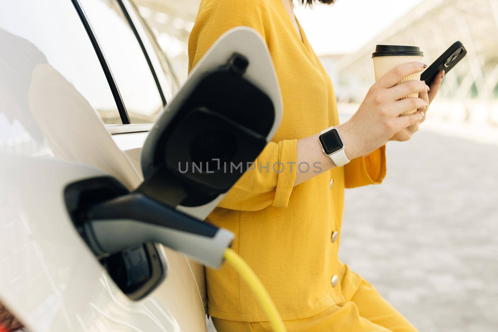 Unrecognizable woman using smart phone and waiting power supply connect to electric vehicles for charging the battery in car. Plug charging an Electric car from charging station by uflypro