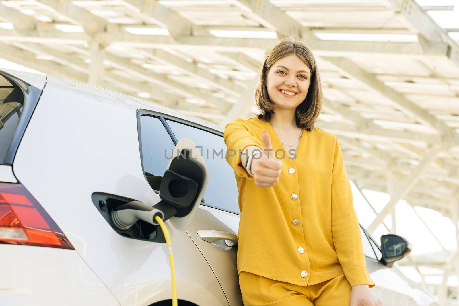 Woman standing on charging station looking at camera and showing thumb up. Businesswoman near electric vehicle outdoors. Eco-friendly modern green transport. Ecology and urban lifestyle