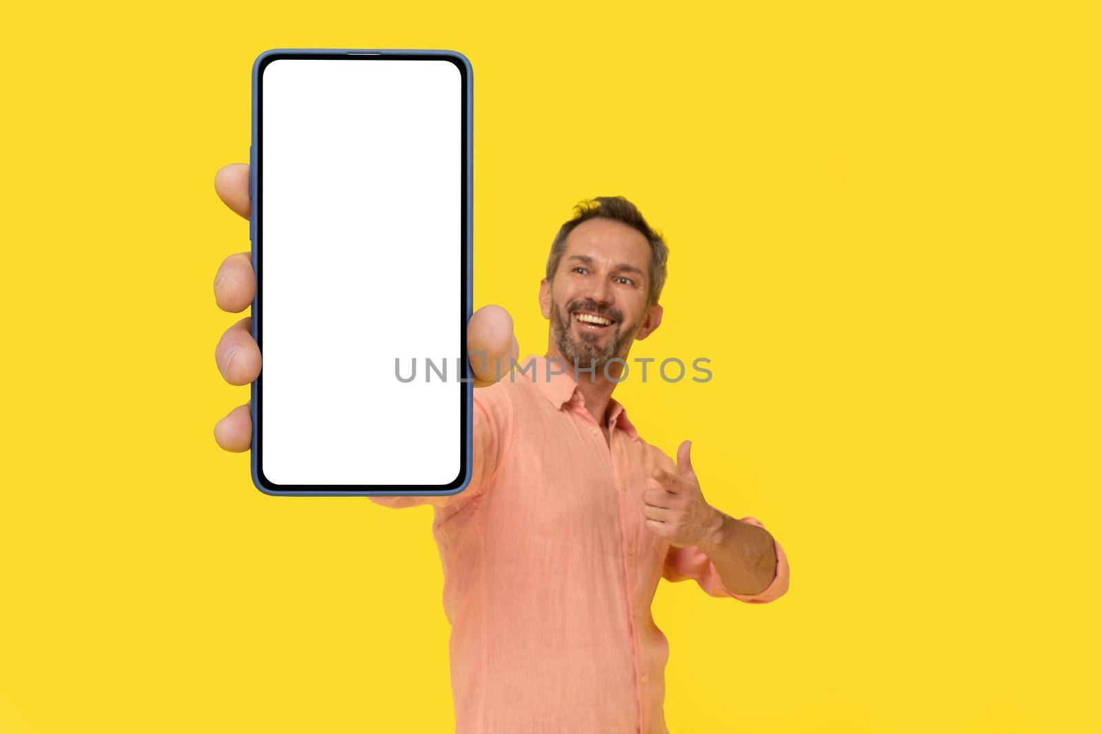 Middle aged grey haired man pointing at smartphone in hand amazed smiling looking at smartphone wearing peach shirt isolated on yellow. Mature fit man with phone, mobile app advertisement.