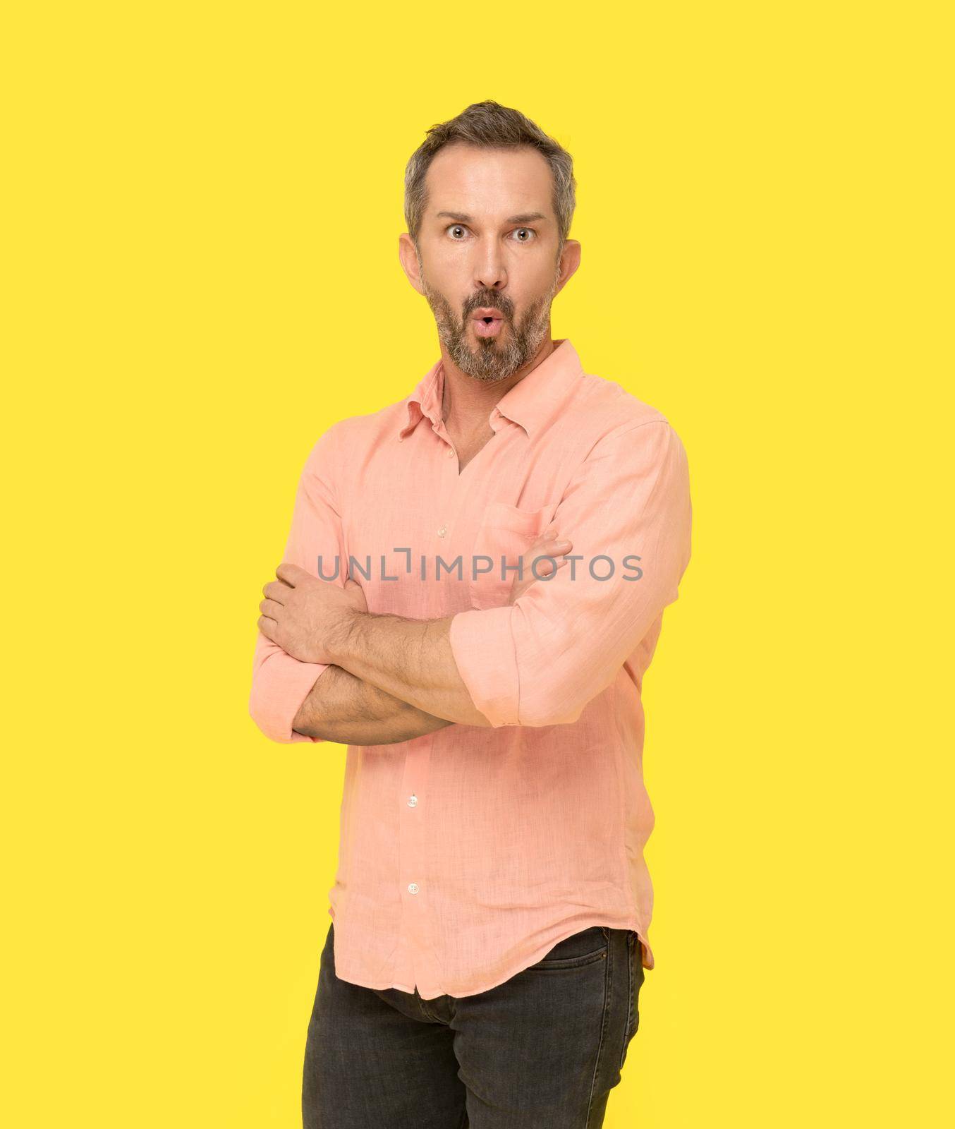 Amazed facial expression middle aged grey haired man standing with arms folded happy looking at camera wearing pink shirt isolated on yellow background. Mature fit man, healthy lifestyle concept.