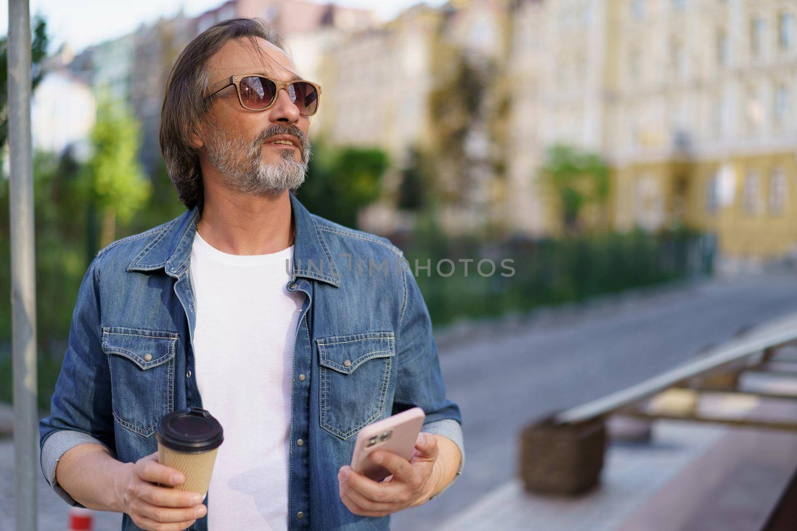 Middle aged man got distracted from mobile phone enjoying coffee outdoors looking at sunset. Middle aged man looking away at street while enjoying free time off work. Business on the go.